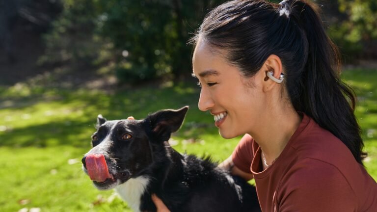 A young woman with dark hair in a ponytail, a dark red top and an Shokz OpenDots ONE earbud in her left ear cuddles a dog. They are in what appears to be a garden on a sunny day and the dog is licking its own nose.