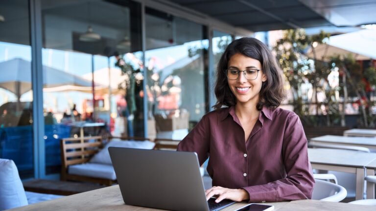 A woman sitting outside in a public space and using a laptop while seated at one of the tables